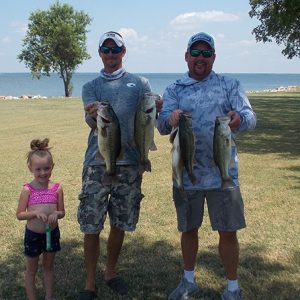 2 men in shorts, tees holding 4 bass in front of Lake Tawakoni