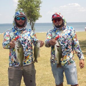 2 men in print hoodies and sunglasses hold 3 bass in front of Lake Tawakoni