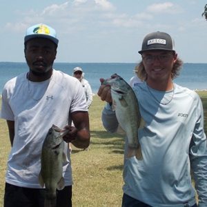2 men in white tees and caps hold 2 bass in front of Lake Tawakoni