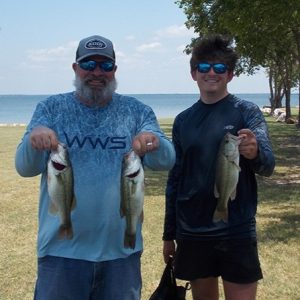 2 men: 1 with grey beard in blue shirt and 1 in black shirt hold 3 bass in front of Lake Tawakoni
