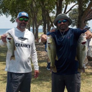man in white shirt and cap with man in black shirt hold up 3 large bass in front of Lake TAwakoni