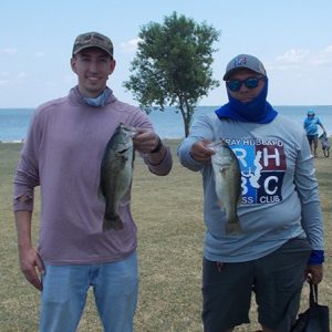 2 men in caps and tees hold 2 bass in front of Lake Tawakoni