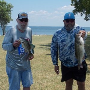 man with white beard in hat holds 1 bass, and man in long sleeve tee holds larger bass in front of Lake Tawakoni