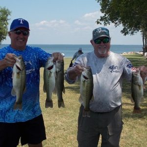 2 men in caps and tees hold up 5 bass in front of Lake Tawakoni