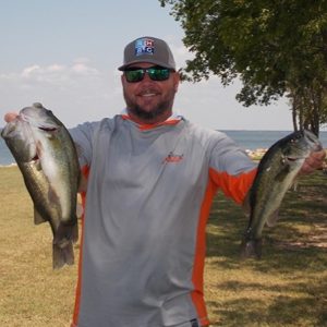 bearded man in white and orange tee holds up 2 large bass in front of Lake Tawakoni