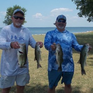 2 men in hats, sunglasses, longsleeve tees holding 4 bass in front of Lake Tawakoni