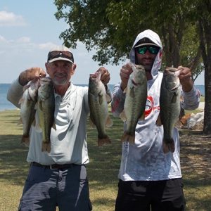 2 men in tees and caps holding 5 bass in front of Lake Tawakoni
