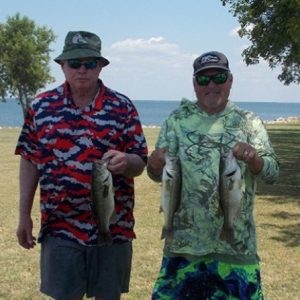 2 men wearing print shirts and caps hold 3 bass in front of Lake Tawakoni