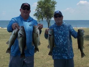 2 men in caps and tees hold up 4 large bass in front of Lake Tawakoni