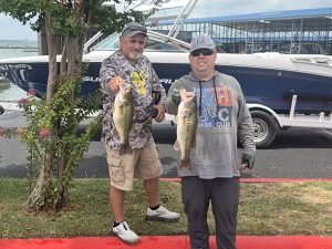 man in camo shirt, cap and tan shorts holds 1 fish, man in grey shirt and cap holds 1 fish in front of lake and boat