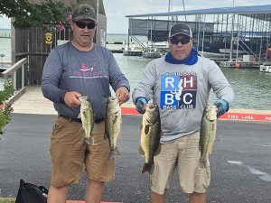 2 men in tshirts and caps hold 2 bass fish each in front of lake