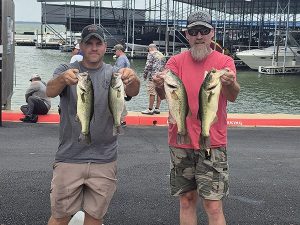 man in grey shirt and hat holds 2 fish, man with long beard in pink shirt holds 2 fish at lake