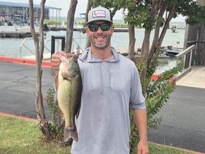 bearded man in grey shirt and cap holds large bass in front of lake