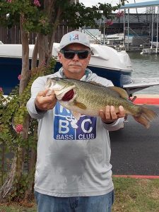 man with sunglasses and white mustache shows of bass fish in front of lake and boat