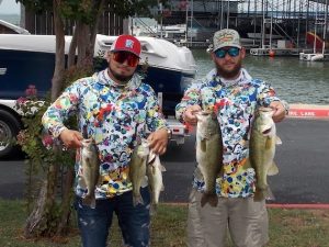 2 men in bright print shirts hold up 4 fish in front of boat at lake