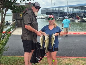 tall bearded man in dark shirt and shorts holds 3 fish with short blonde woman in blue shirt and black shorts