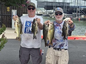 2 men at lake holding fish, wearing tshirts, ball caps and sunglasses