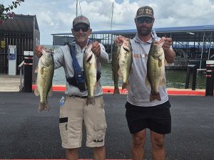 2 men in grey tshirts and shorts hold 2 fish in front of lake